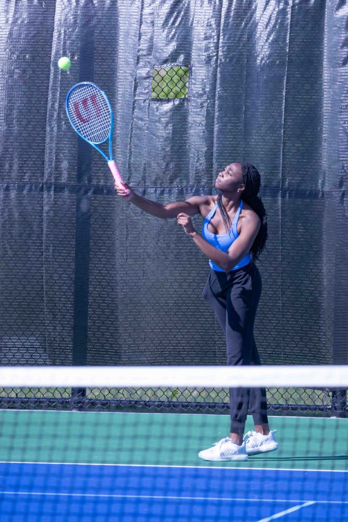 A young woman in a blue athletic top and black pants prepares to serve a tennis ball on a court.