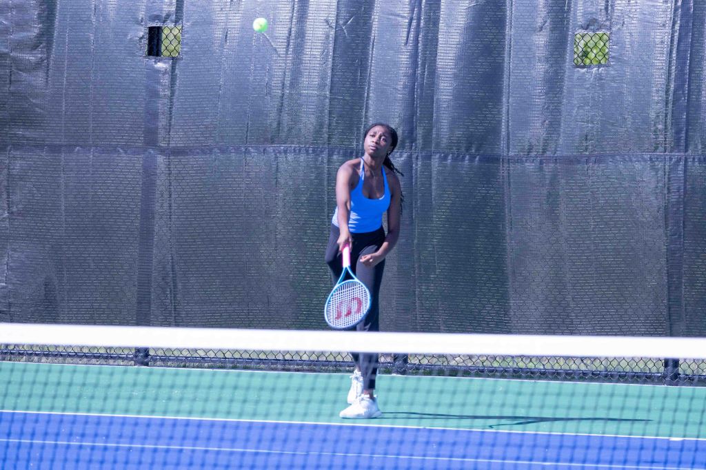A young woman in a blue tank top and black leggings prepares to hit a tennis ball on a court, with a net in the foreground.