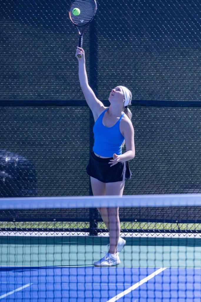 A young woman serving a tennis ball on a court, wearing a blue tank top and a black skirt, with a focused expression.