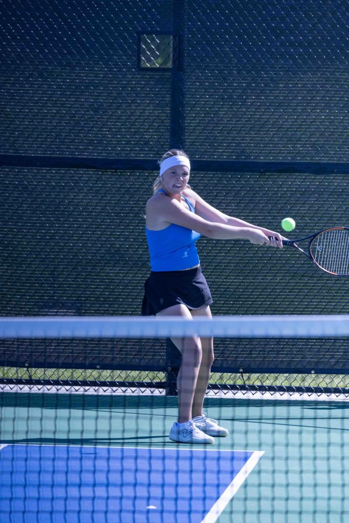 A female tennis player in a blue tank top and black skirt is mid-swing, preparing to hit a tennis ball on a court with a fence in the background.