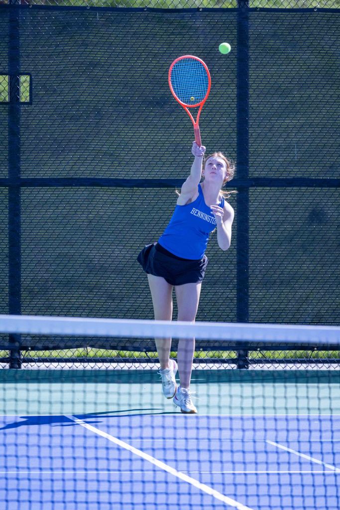 A female tennis player serving the ball on an outdoor court, wearing a blue tank top with 'Bennington' on it and a black skirt.