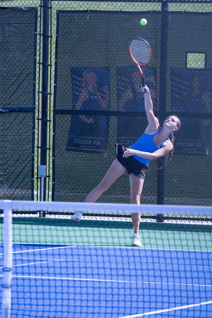 A young woman in a blue tank top and black shorts serves a tennis ball on a court, with a net in the foreground and a green backdrop.