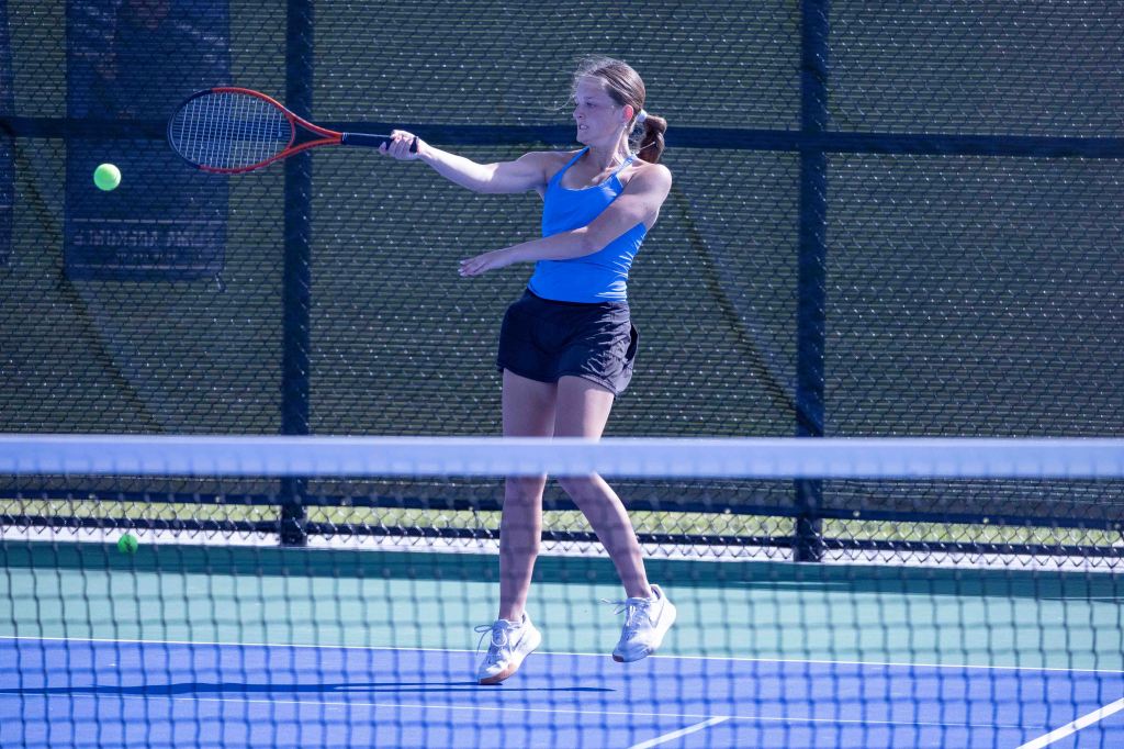 A female tennis player in a blue tank top and black shorts is hitting a tennis ball with a racket on a blue court.