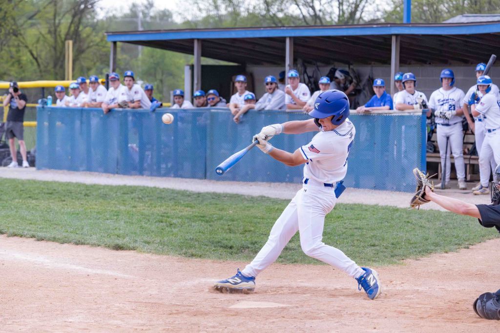 A young baseball player in a white uniform swings a bat to hit a baseball during a game, with teammates watching from the dugout in the background.