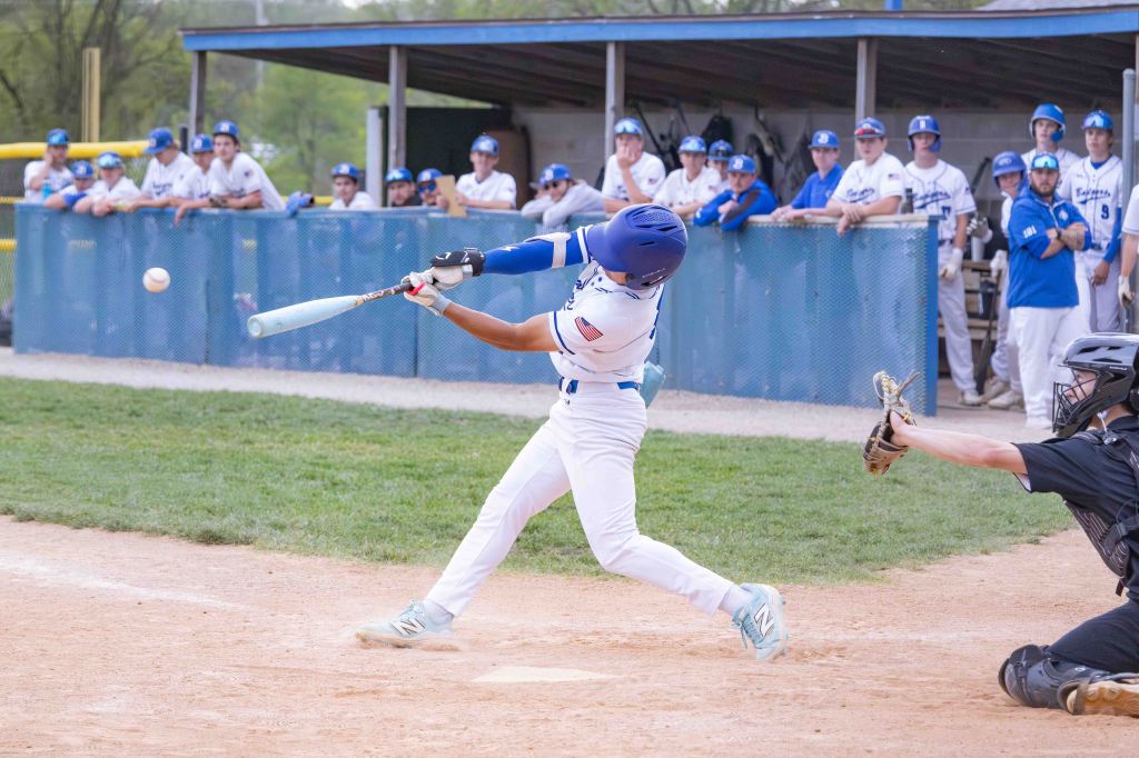 A baseball player swings at a pitch during a game, with a catcher ready to receive the ball and teammates watching from the dugout.
