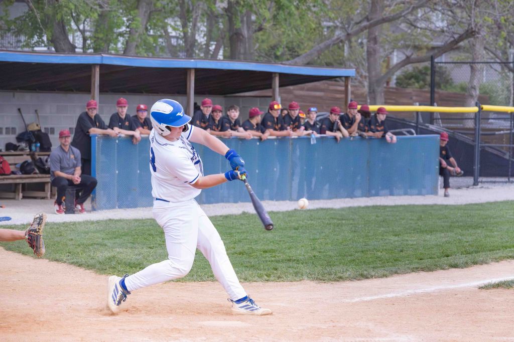 A young baseball player swinging a bat at a pitch during a game, with teammates in the background watching from the dugout.
