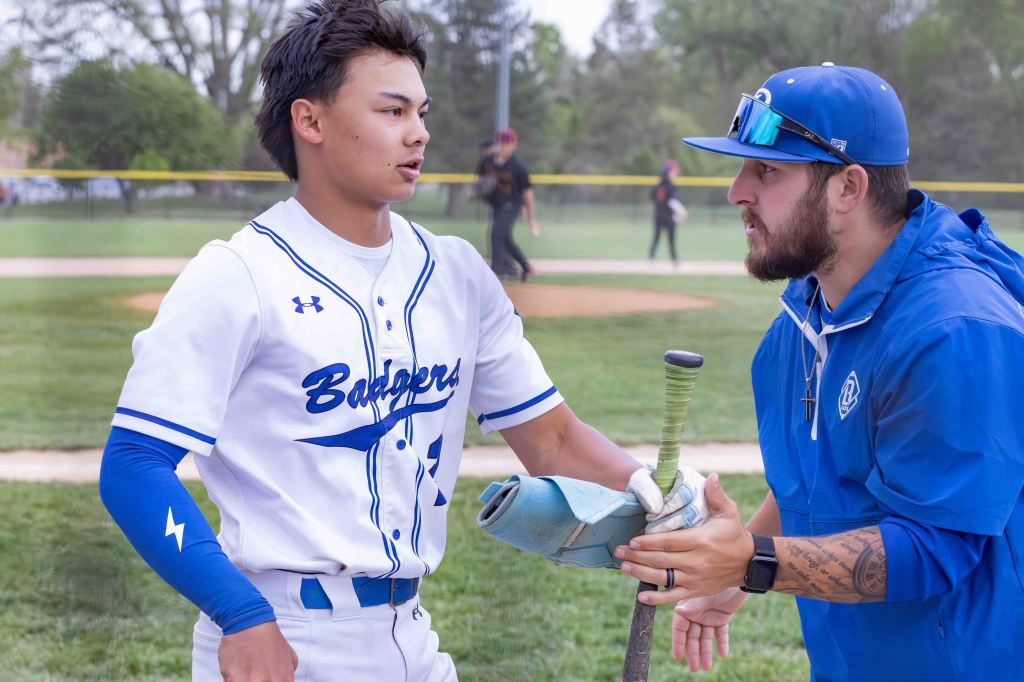 A baseball player in a white uniform with blue accents talks to a coach wearing a blue jacket, both engaged in a focused discussion on a baseball field.