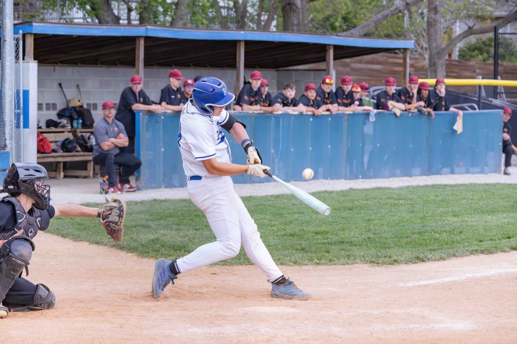 A baseball player in a white uniform swings a bat to hit a ball, with a catcher in black gear crouched behind home plate and a team of players observing from the dugout in the background.