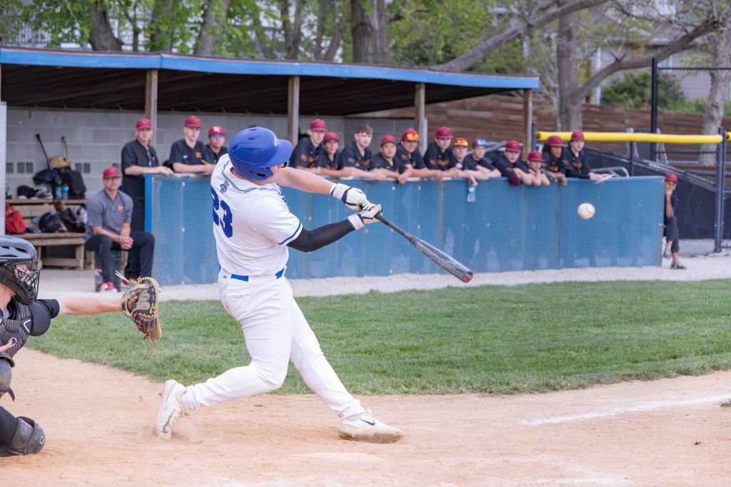 A baseball player in a white uniform swings a bat at a flying baseball, while a catcher in black gear crouches behind home plate. A group of players in red hats watches from the dugout in the background.