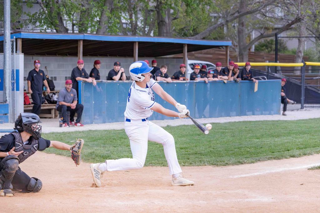 A young baseball player at bat swings the bat, making contact with the ball. In the background, teammates in uniforms observe from the dugout.