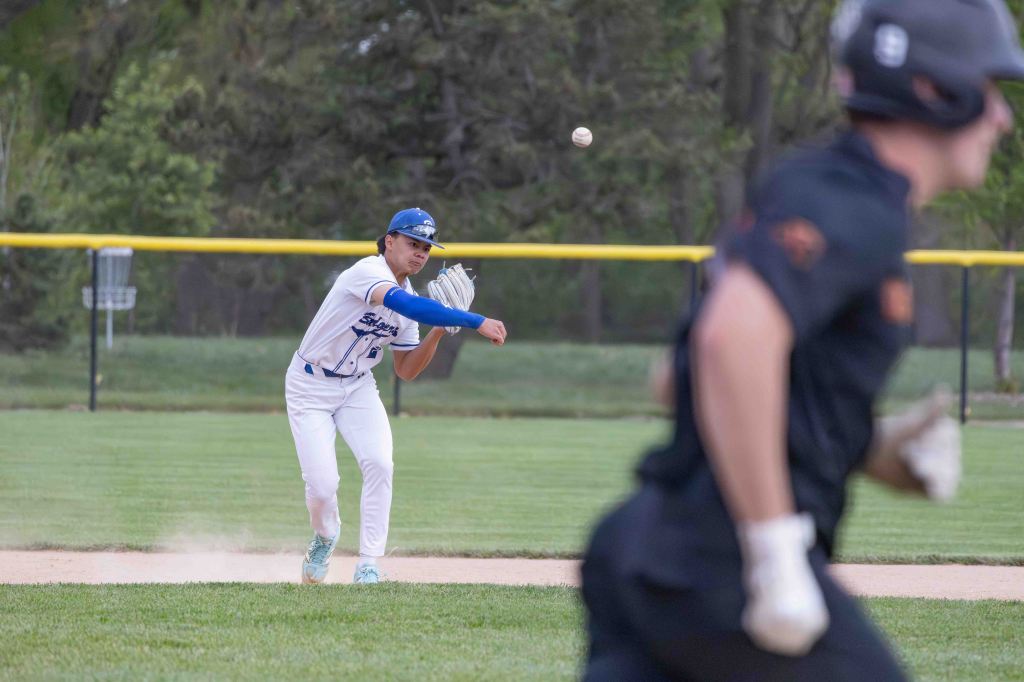 A baseball player in a white uniform is throwing the ball while another player in a dark uniform is running past him on the base path.