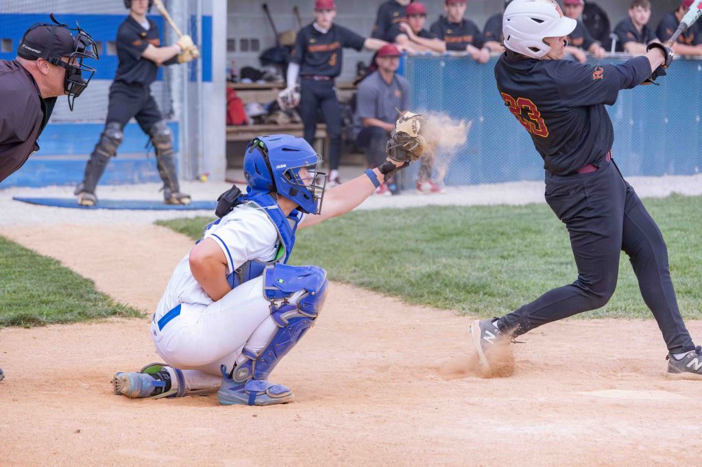 A baseball game scene showing a catcher in blue gear squatting behind home plate, an umpire in black behind him, and a batter in a black jersey swinging at a ball, with dust in the air from the impact.