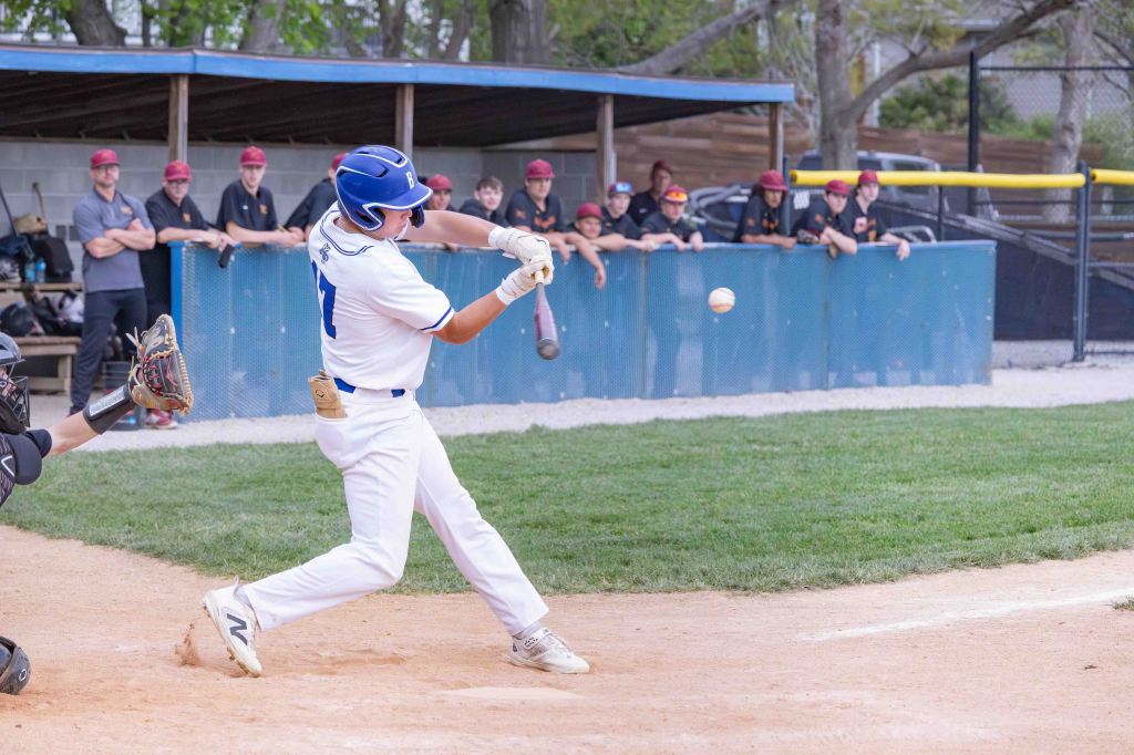 A baseball player swings at a pitch during a game, with teammates watching from the dugout in the background.