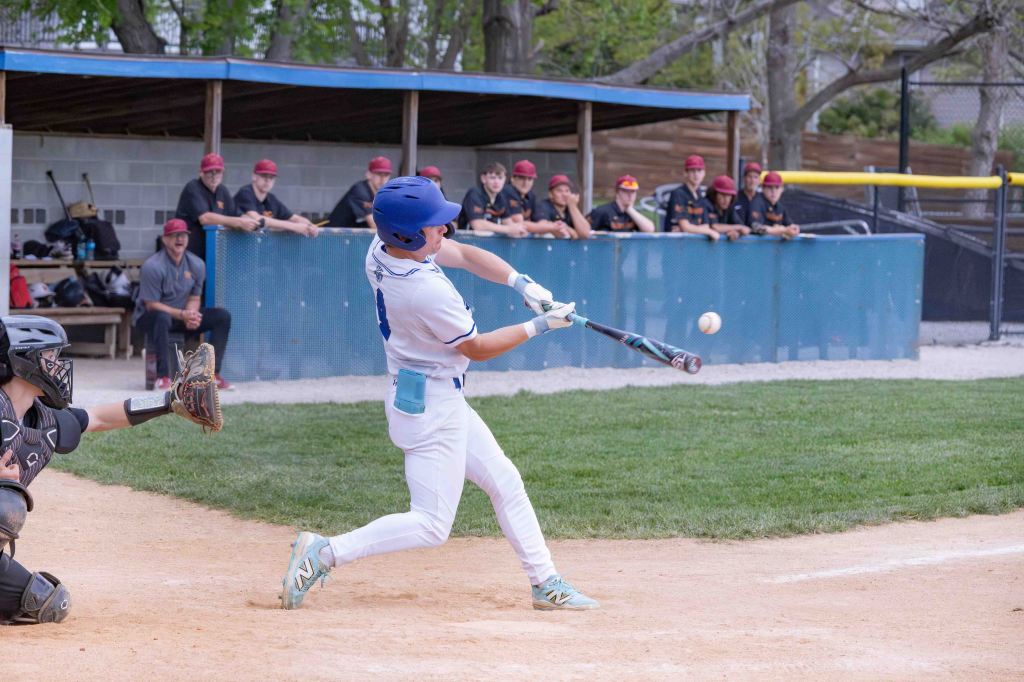A baseball player in a white uniform swings a bat to hit a ball while a catcher in black gear is crouched behind home plate. In the background, a group of players in dark uniforms watches from the dugout.