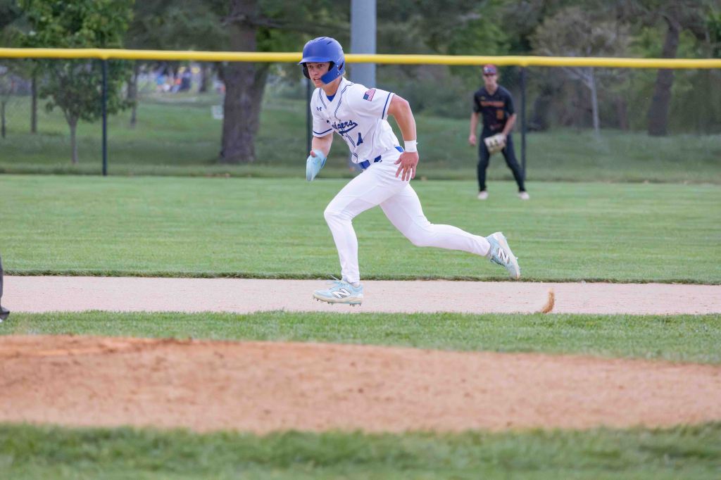 A baseball player in a blue helmet and white uniform sprints down the base path during a game, with a fielder in black shorts standing in the background.