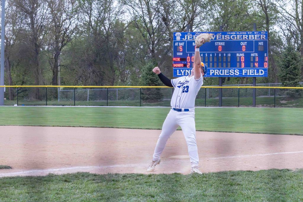 A baseball player in a white uniform throws a ball from the infield toward a scoreboard, displaying the game scores and statistics, at a baseball field.