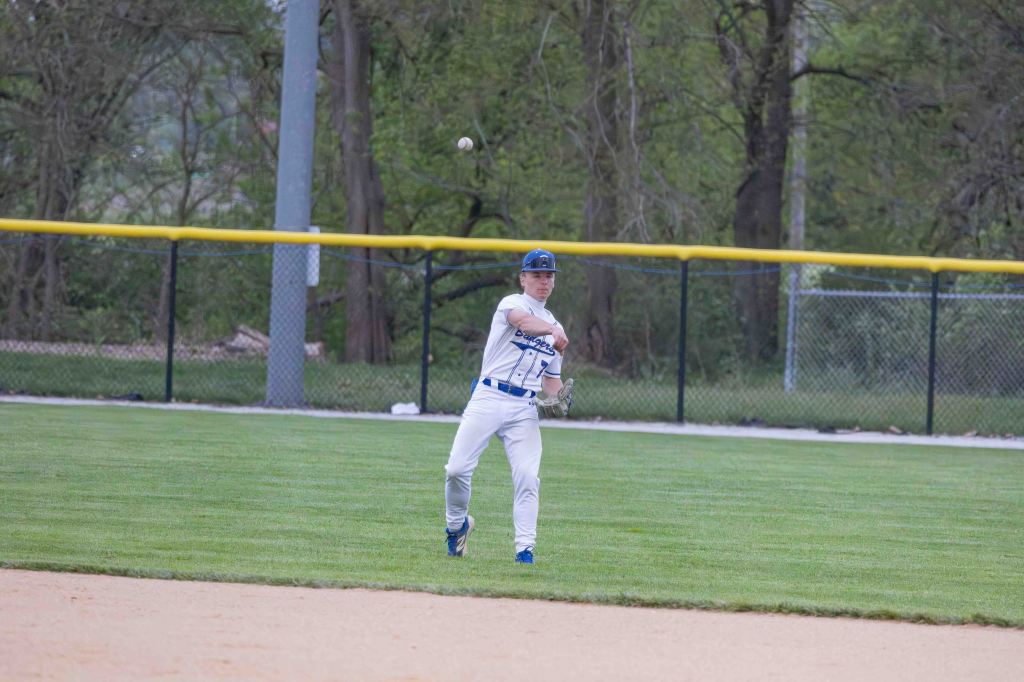A young baseball player in a white and blue uniform throwing a baseball from the outfield on a grassy field.
