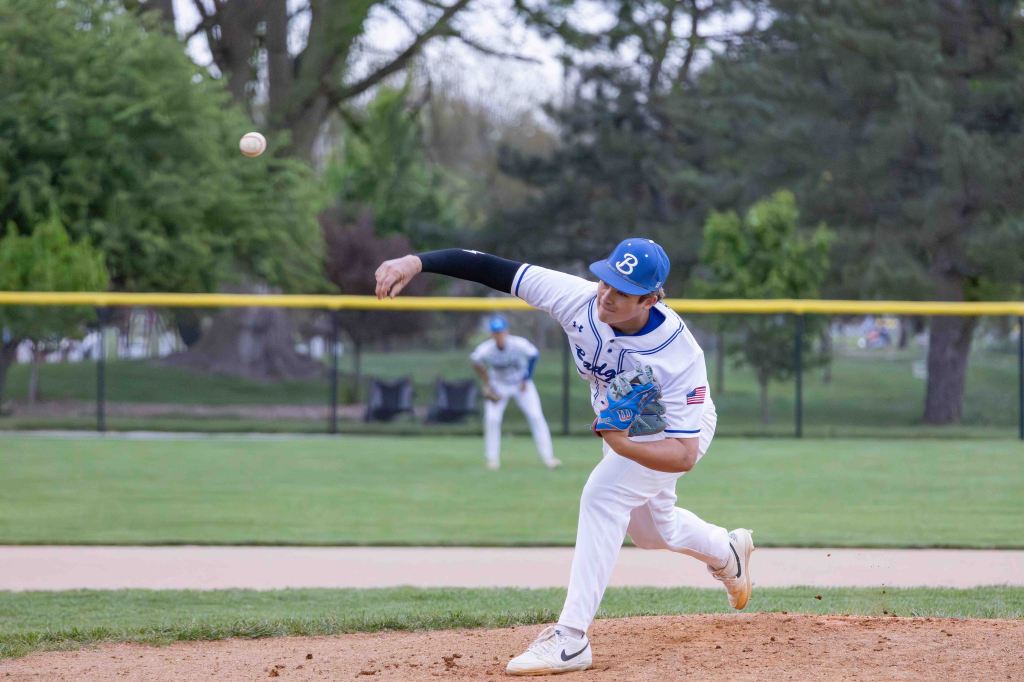 A young baseball pitcher in a blue and white uniform is in the process of throwing a baseball on a field. Another player stands in the background.