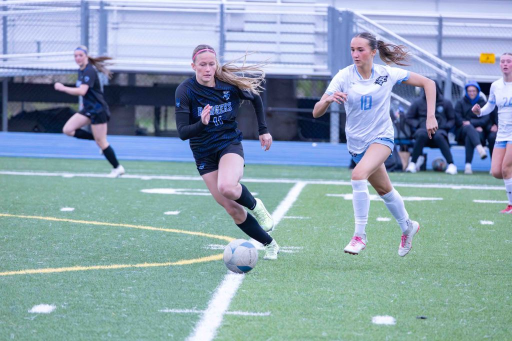 Two female soccer players sprinting on the field, one in a black jersey with the number 11 dribbling a soccer ball, while the other in a white jersey with the number 10 is closely following.
