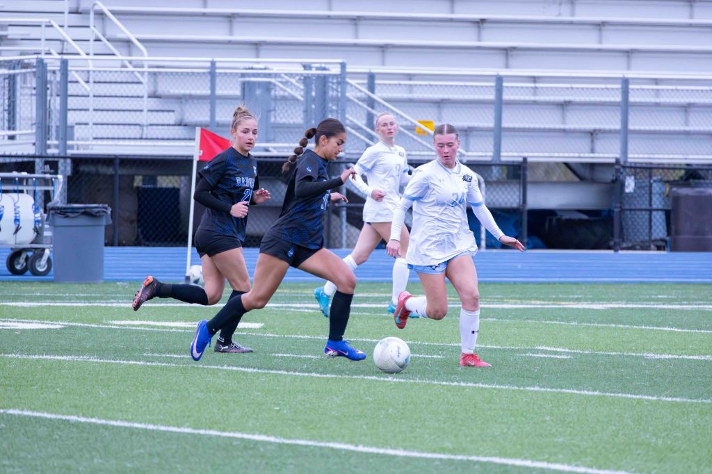 A soccer game in progress with four female players, two in black jerseys and two in white jerseys, competing for the ball on a grassy field.