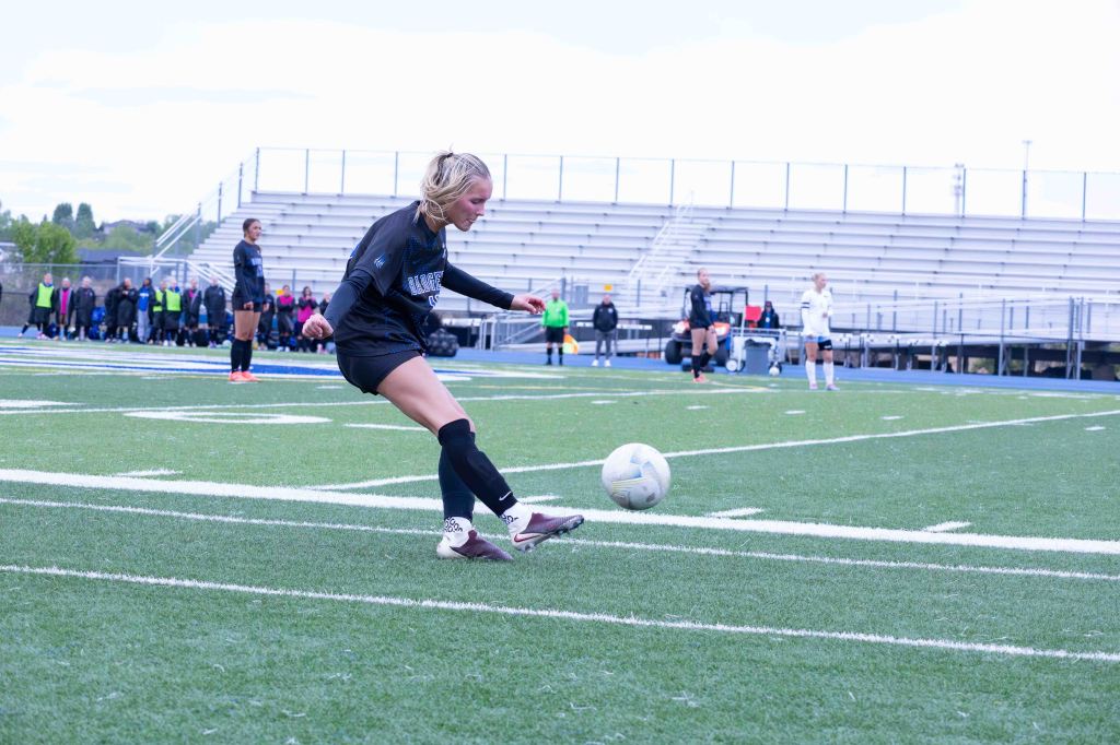 A female soccer player wearing a black jersey and shorts is kicking a soccer ball on a grassy field, with teammates and spectators in the background.