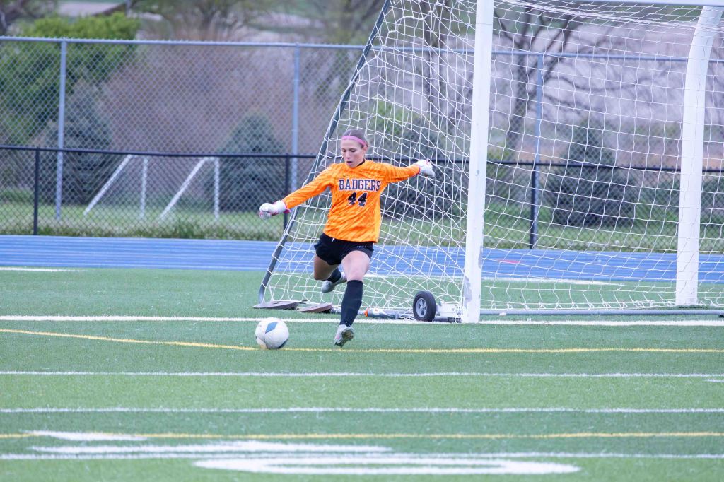 A female soccer goalkeeper in an orange uniform is preparing to kick a soccer ball on a field with a goal in the background.