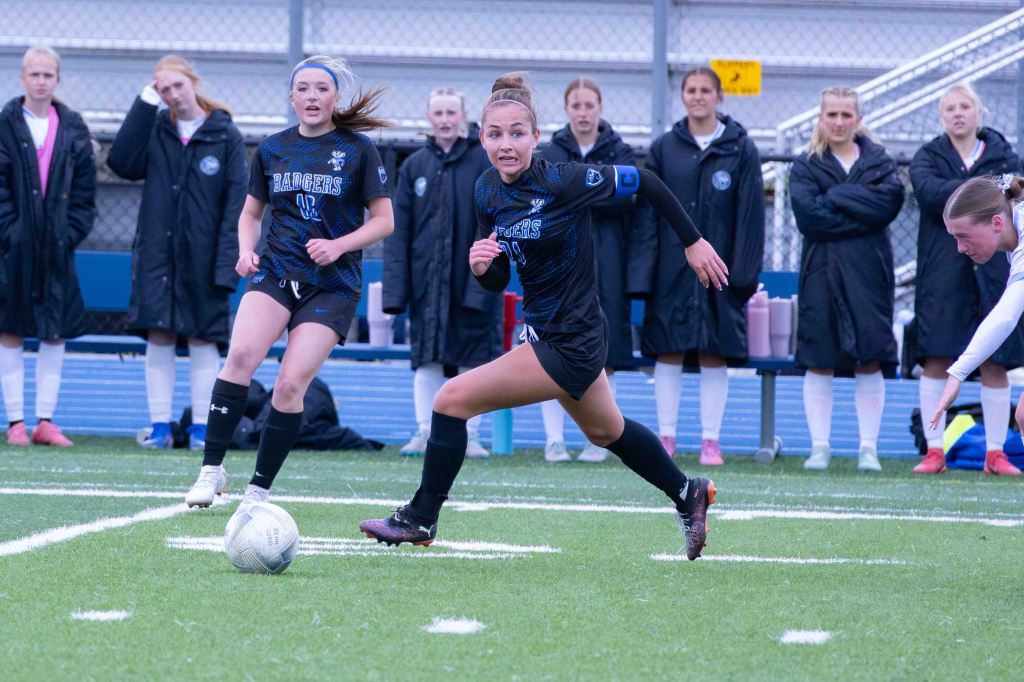 A soccer player in a black uniform sprinting with the ball during a game, while teammates and spectators in the background watch. The player wears a captain's armband.