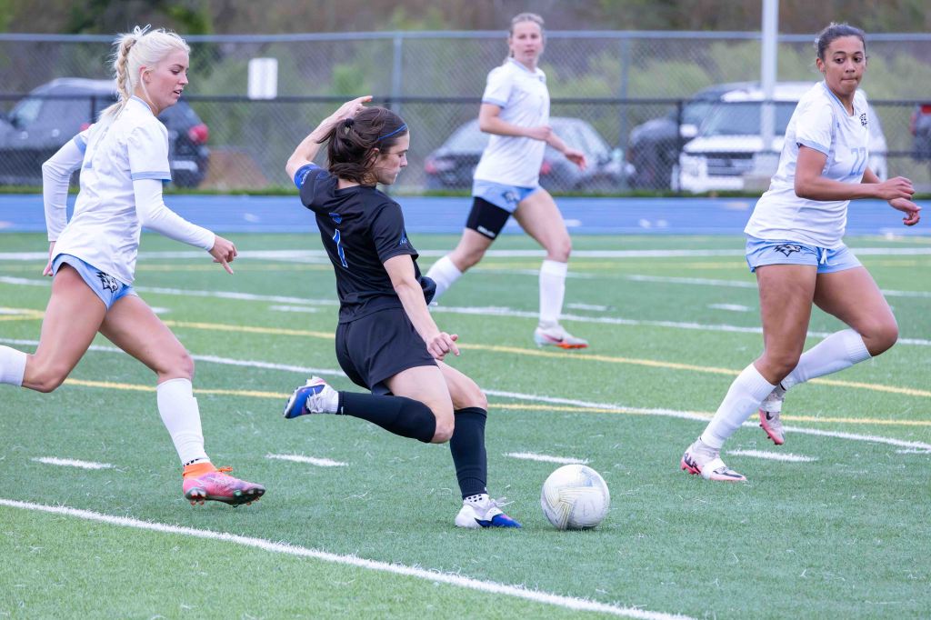 A soccer match in progress with a player in a black jersey kicking the ball while two defenders in white jerseys attempt to block the play. A third player in the background is monitoring the action.