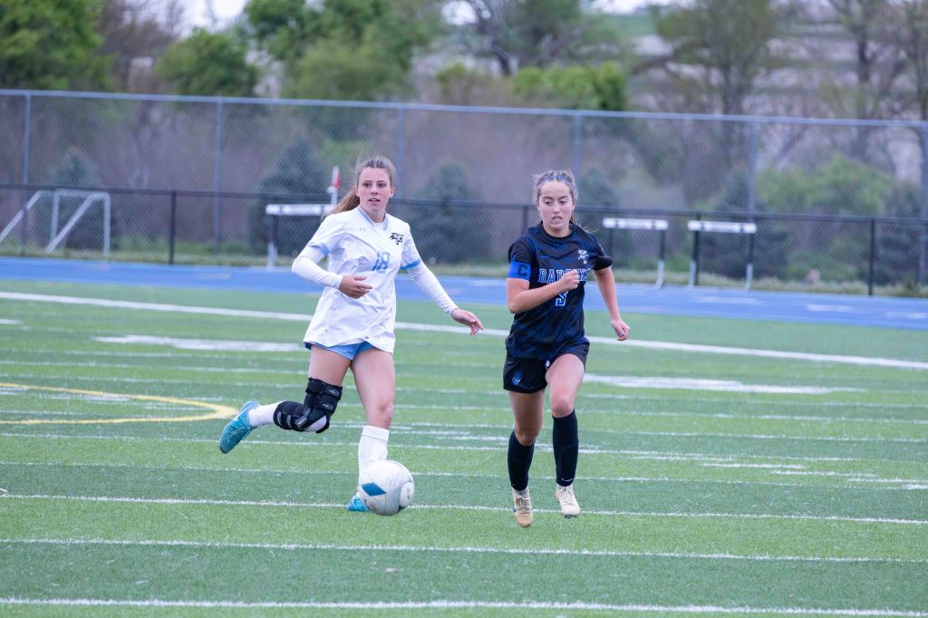 Two female soccer players compete for the ball on a green turf field. One player is wearing a white jersey with the number 18, while the other is in a black jersey with a captain's armband.