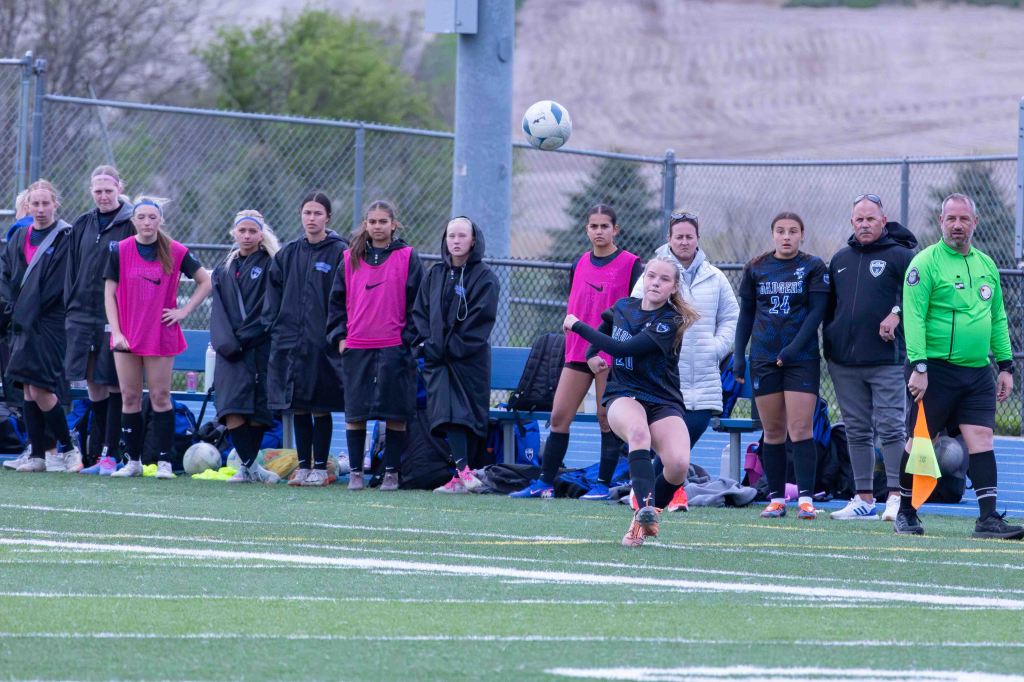 A female soccer player in a black uniform performs a throw-in while teammates and coaches watch from the sidelines. Several players are dressed in pink training vests, and the scene captures the energy of a game in progress.