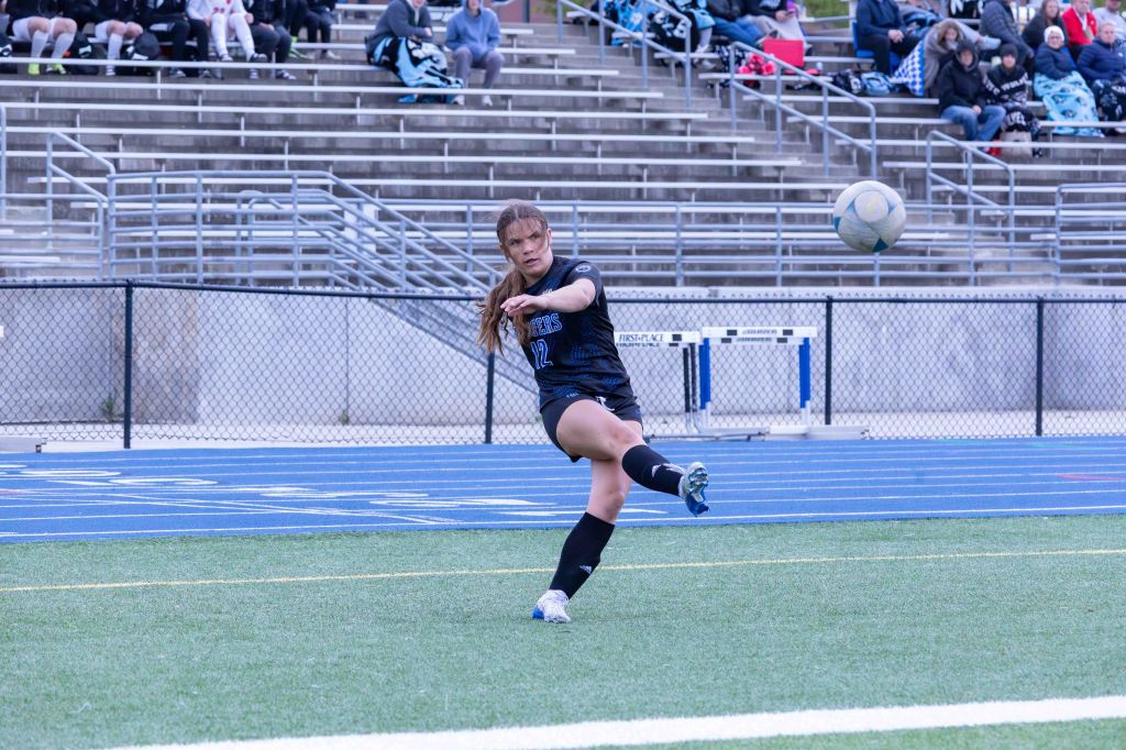 A female soccer player in a black uniform, number 12, executing a kick on the field with a ball in mid-air, while spectators watch from the stands.