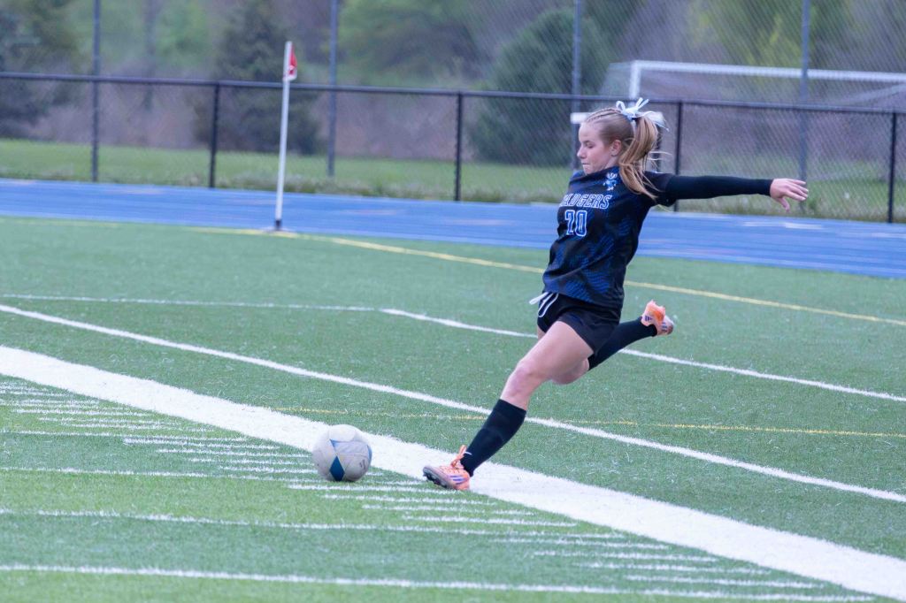 A female soccer player taking a corner kick on a field, wearing a blue and black jersey with the number 20. A soccer ball is positioned on the ground near her foot.