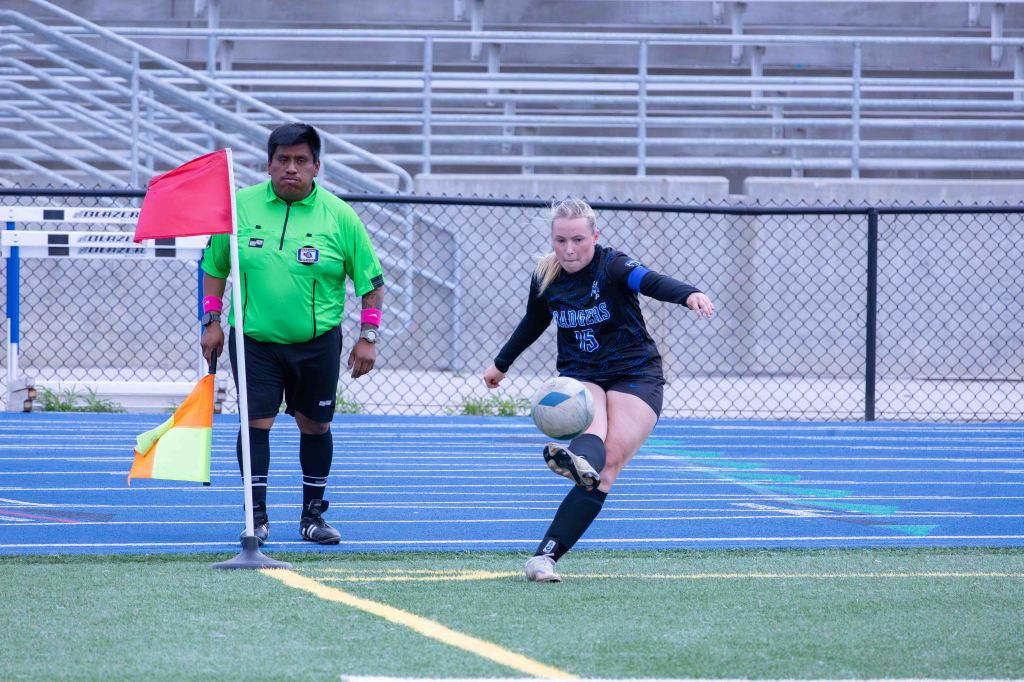 A female soccer player in a black jersey and shorts kicking a soccer ball on the field, with a referee in a green uniform and flags nearby.