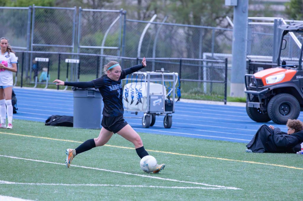 A high school female soccer player in a black uniform takes a shot at the ball on a grass field, with a blue track in the background and another player watching.