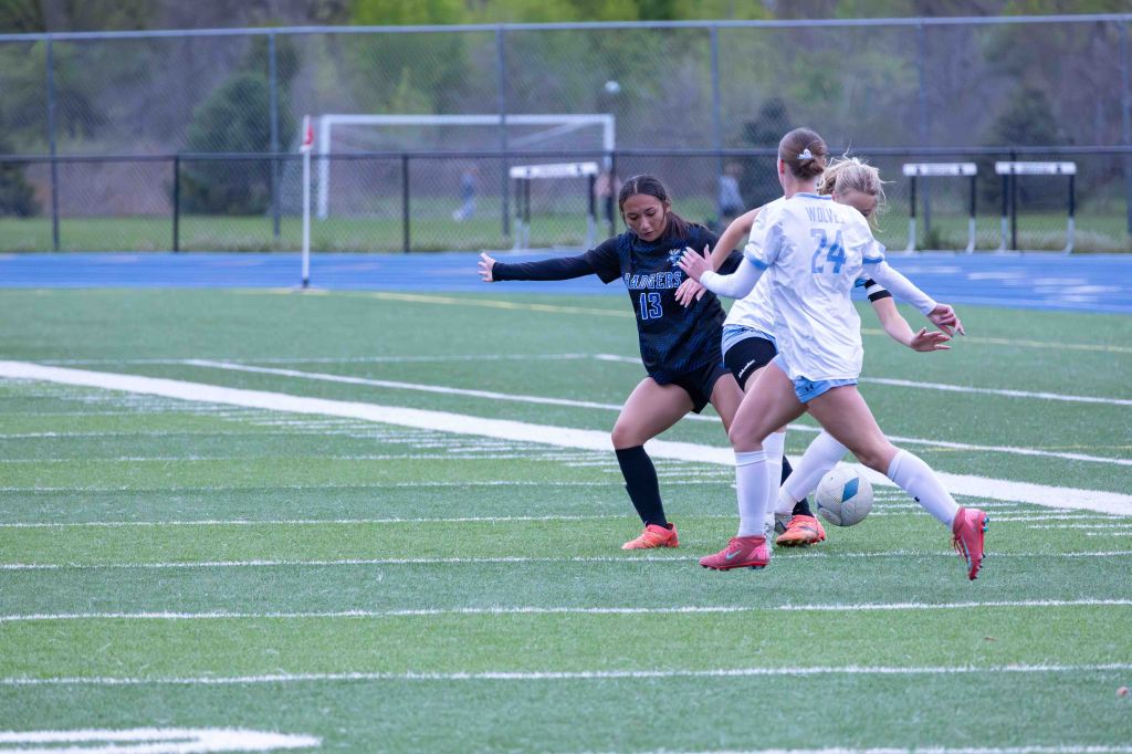 A soccer match scene showing two players competing for the ball on a grassy field, with one player in a black jersey and another in a white jersey.