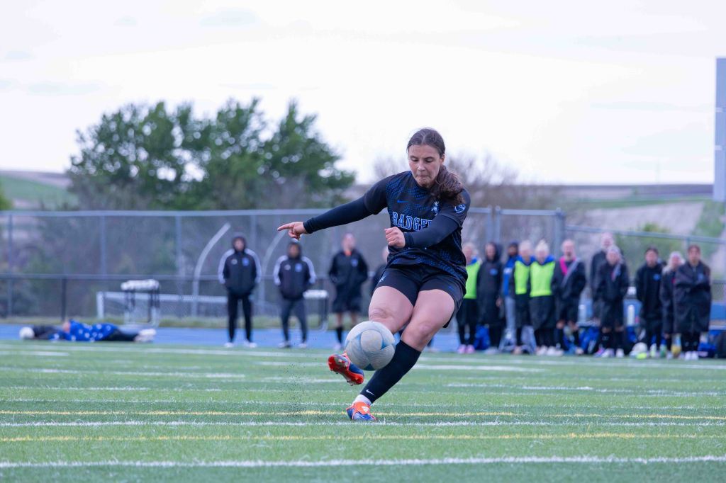 A female soccer player kicks a soccer ball on the field during a game, with spectators in the background.
