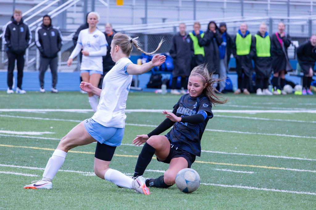 Two female soccer players are engaged in a play on the field, one sliding on the ground and reaching for the ball while the other stands nearby.