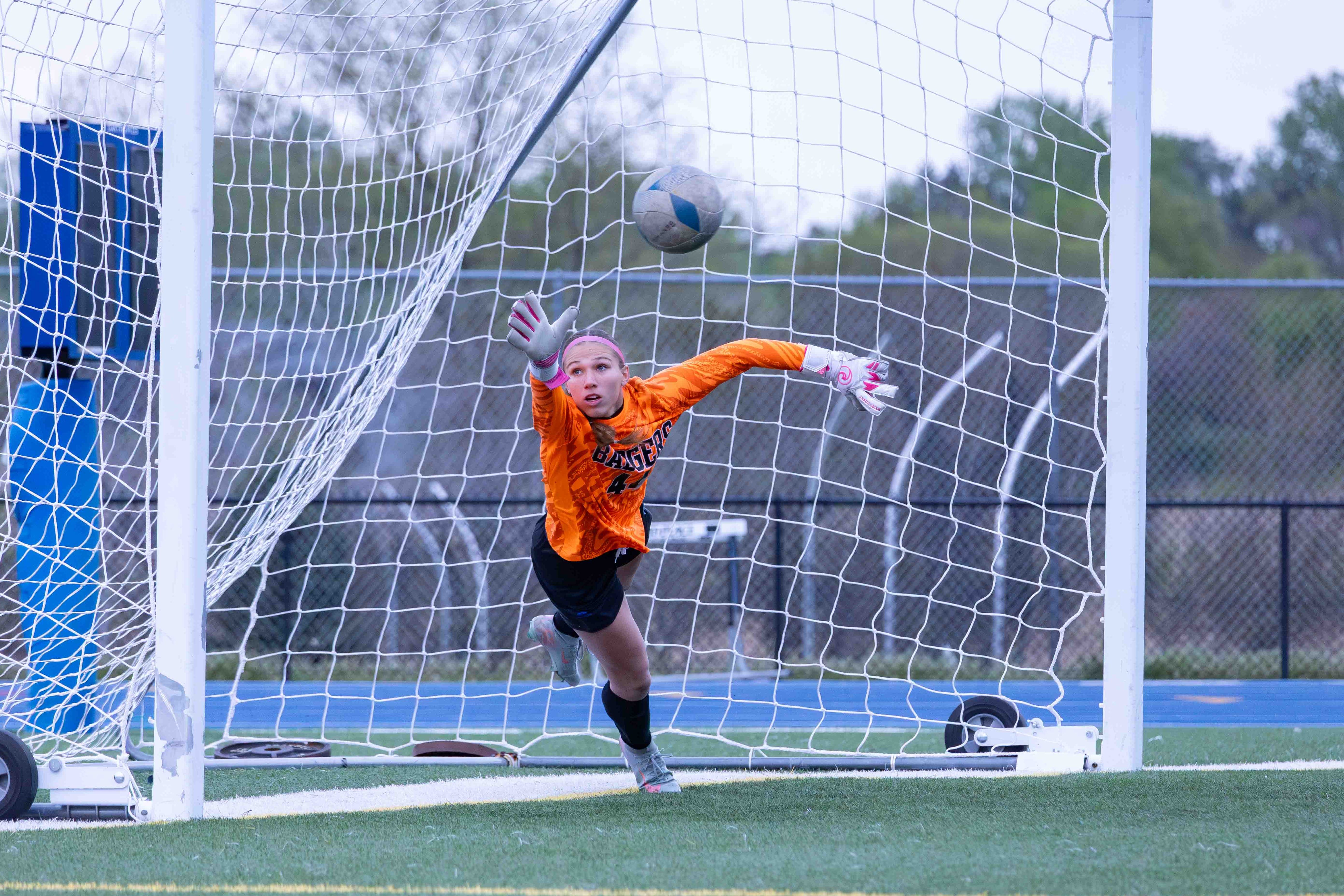 A young female soccer goalkeeper in an orange jersey dives to save a soccer ball during a game, with the goal net in the foreground.