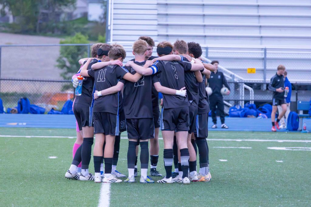 A group of young male soccer players in black jerseys huddle together on a green field, showing teamwork and camaraderie before a game.