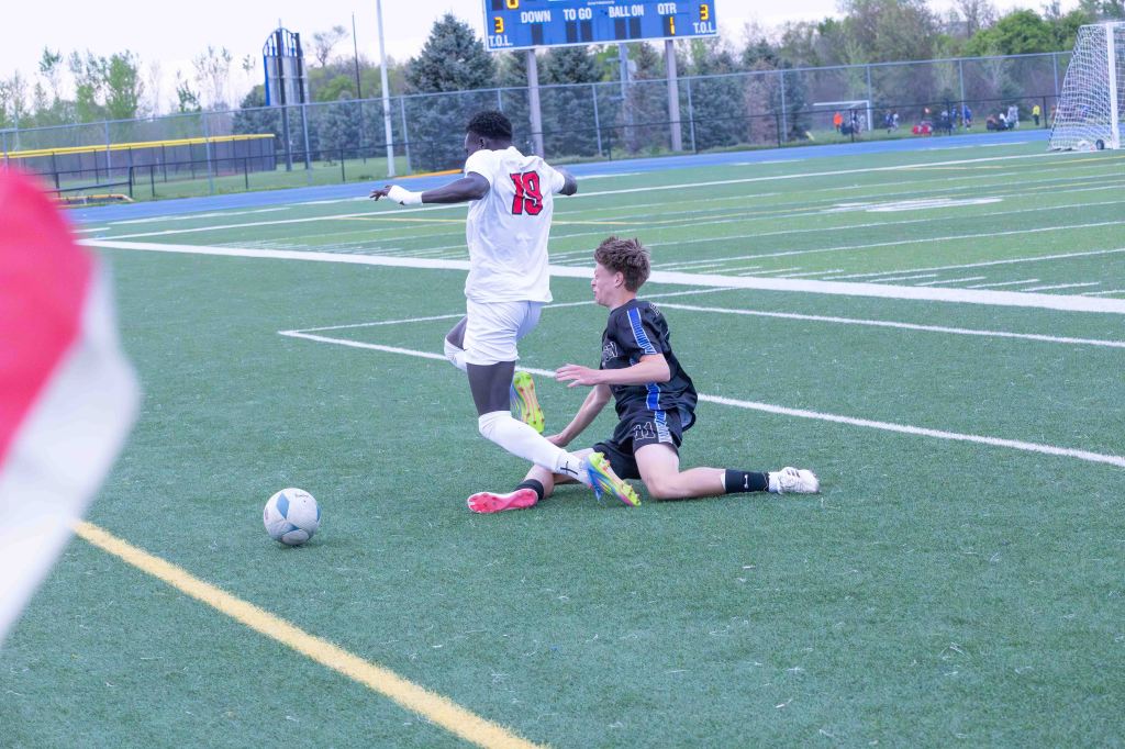 A soccer match scene showing two players competing for the ball; one player in a white jersey with the number 19 falls while the other player in a black jersey with the number 21 assists in the play.