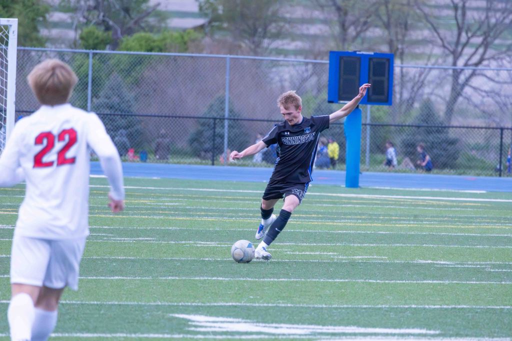 A young male soccer player in a black jersey is preparing to kick a soccer ball on a grass field, while another player in a white jersey with the number 22 is running in the background.