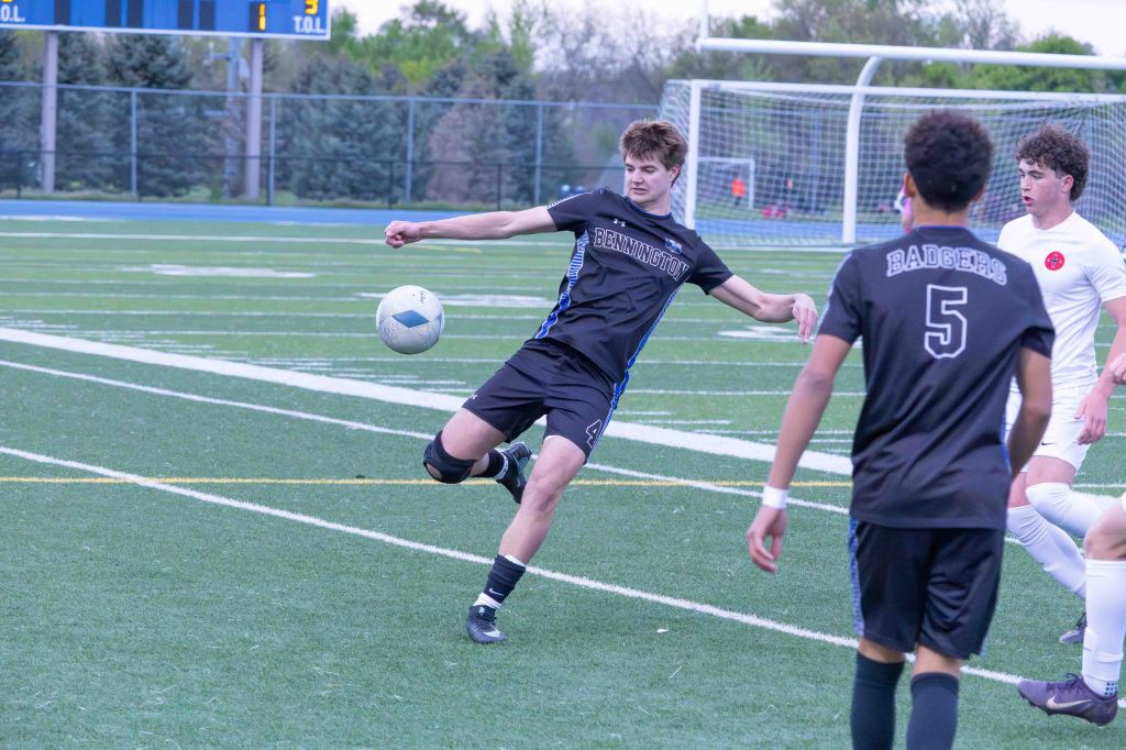 A soccer player in a black jersey with 'Bennington' printed on it is kicking a soccer ball on a grassy field during a game, with other players in the background.