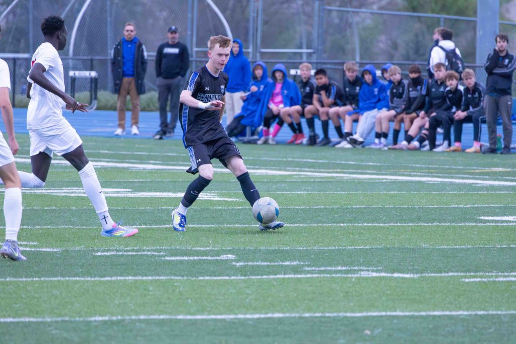 A soccer player in a black uniform dribbles a ball on a field, with an opponent in a white uniform attempting to block him. In the background, several players sit on the sidelines watching the game.