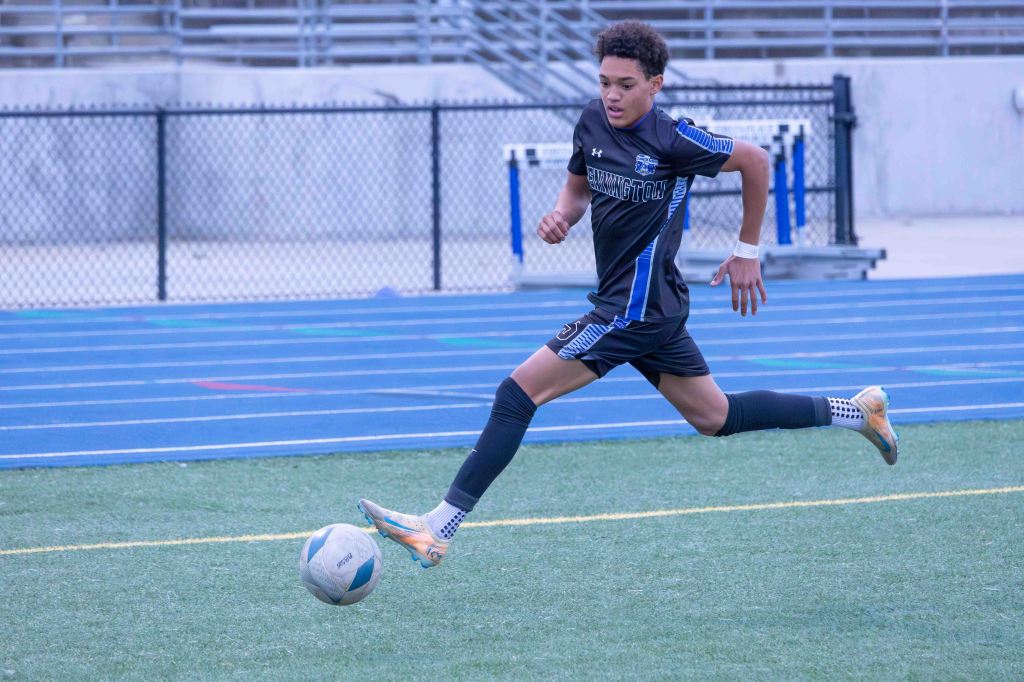 A young male soccer player in a black uniform running while kicking a soccer ball on a field with blue track lines and a fence in the background.