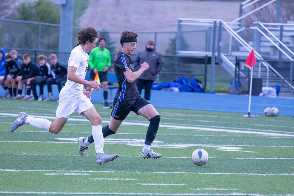 Two young male soccer players competing for the ball on a grass field, with one player in a white jersey and the other in a black jersey. A bench of teammates is visible in the background.