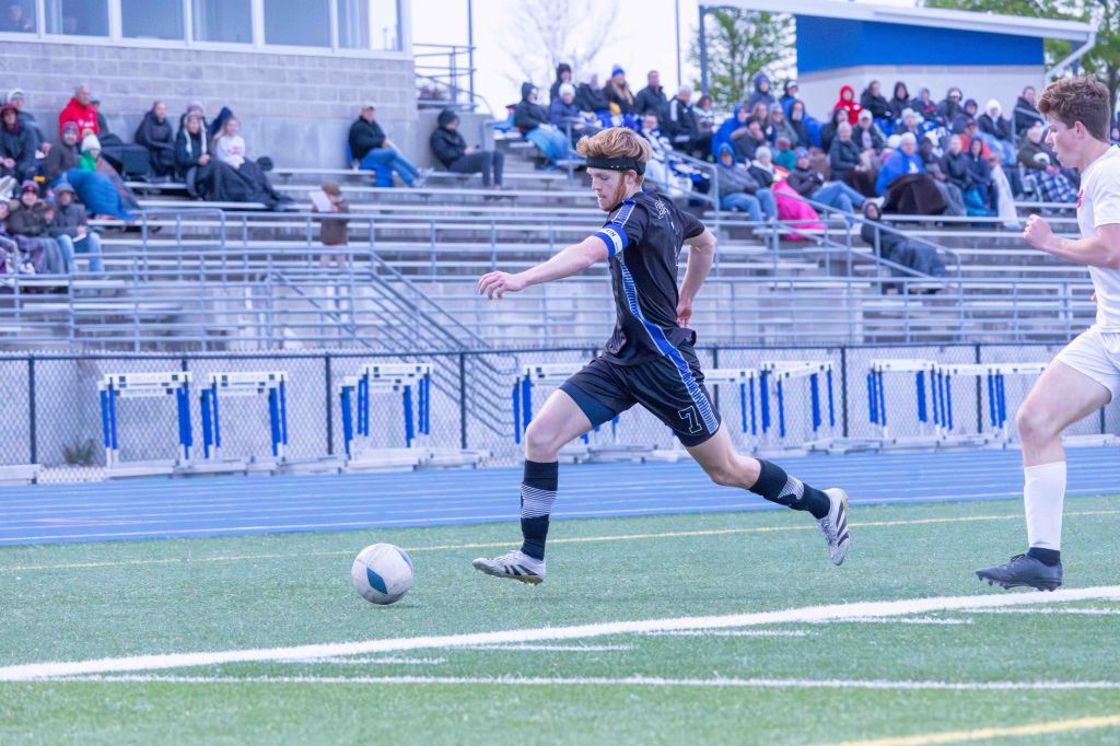 A soccer player in a black jersey with the number 7 chases after a ball on a grass field, while a defender in a white jersey approaches from the right. Spectators are visible in the stands in the background.