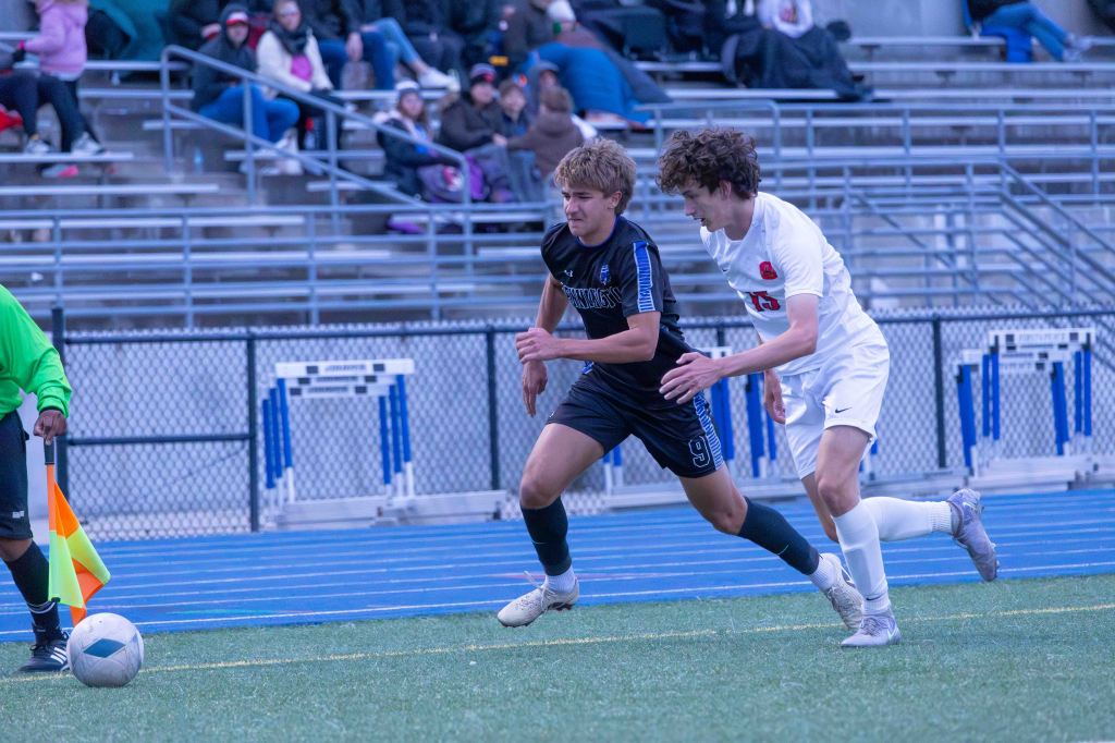 Two high school soccer players compete for the ball on a field, with one player in a blue uniform and the other in a white uniform. A referee is nearby, holding a flag.