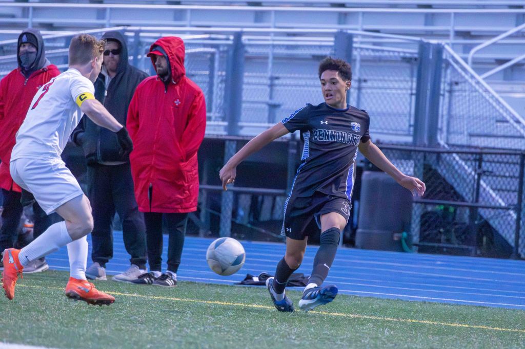 A soccer player in a black jersey with 'Bennington' written on it is skillfully dribbling a ball on the field, while an opposing player in a white jersey watches. Coaches and spectators in red jackets are visible in the background.