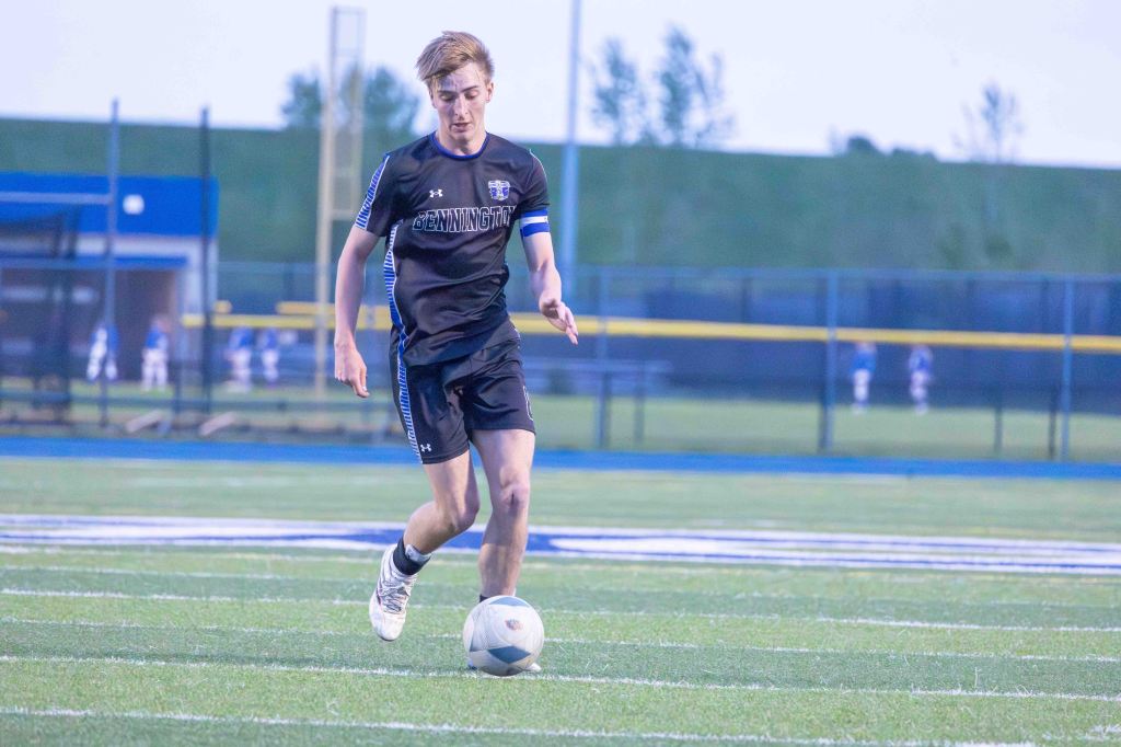 A male soccer player in a black jersey and shorts is dribbling a soccer ball on a grassy field during a game.