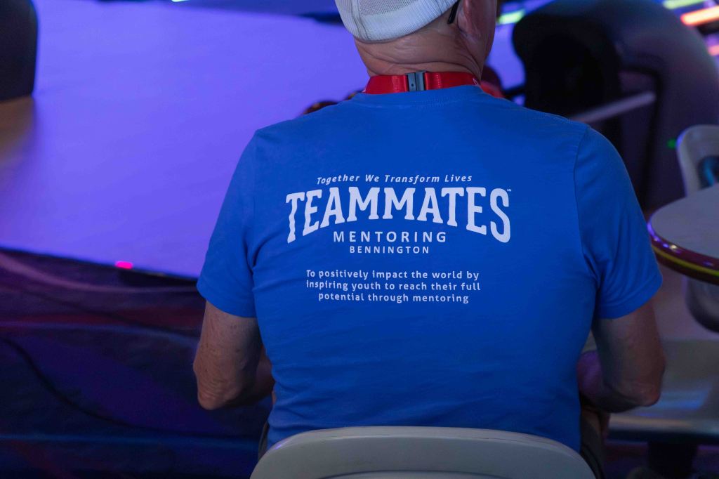 A person sitting with their back to the camera, wearing a blue shirt displaying the text 'TEAMMATES MENTORING BENNINGTON' and a mission statement about positively impacting youth through mentoring.